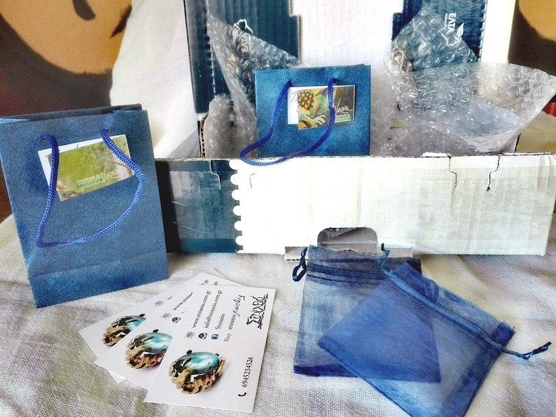 May include: An assortment of packaged jewelry items. The image shows blue gift bags, small blue drawstring pouches, and business cards with a logo and contact information. The items are arranged in a cardboard box with bubble wrap.