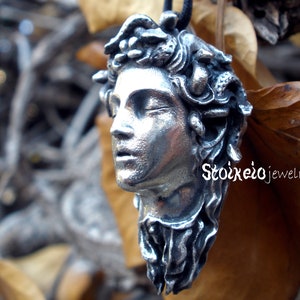 May include: A silver-toned pendant shaped like a human head with snakes for hair, featuring closed eyes and a slightly open mouth. The pendant is suspended from a black cord and displays the text Stoixeiojewelry in white lettering on the side.