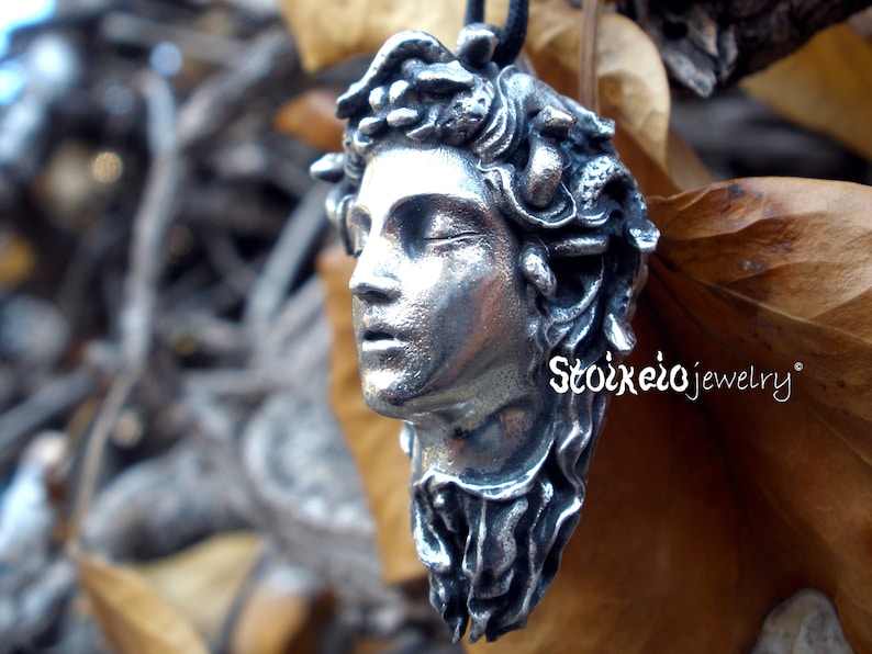 May include: A silver-toned pendant shaped like a human head with snakes for hair, featuring closed eyes and a slightly open mouth. The pendant is suspended from a black cord and displays the text Stoixeiojewelry in white lettering on the side.