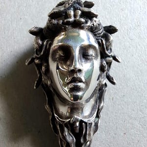 May include: A polished silver pendant featuring a sculpted human face with closed eyes and a serene expression. The head is adorned with intricate, textured snake-like forms that cascade down the sides, creating a detailed, three-dimensional metallic relief.