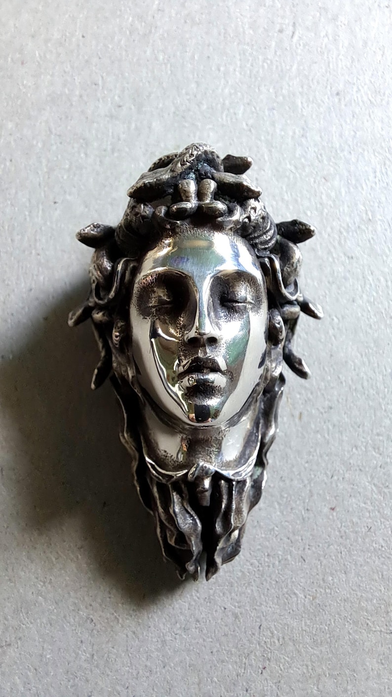 May include: A polished silver pendant featuring a sculpted human face with closed eyes and a serene expression. The head is adorned with intricate, textured snake-like forms that cascade down the sides, creating a detailed, three-dimensional metallic relief.