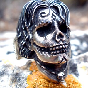 May include: A silver-colored skull ring with intricate details. The skull features stylized hair, facial markings, and a small skull pendant. The ring is set against a natural, textured background.