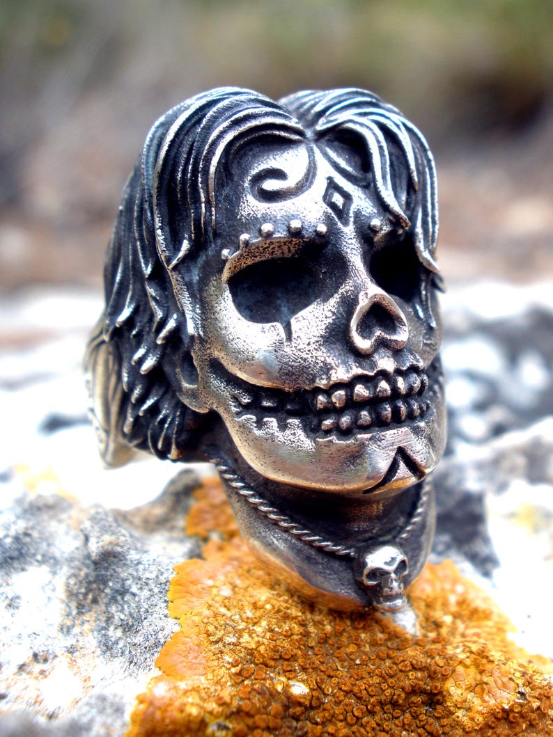 May include: A silver-colored skull ring with intricate details. The skull features stylized hair, facial markings, and a small skull pendant. The ring is set against a natural, textured background.