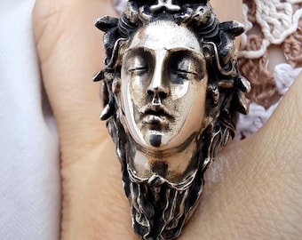 May include: A silver ring featuring a detailed sculptural face with closed eyes and flowing hair. The metal has an oxidized finish that highlights the deep contours and intricate textures of the facial features and the surrounding hair strands.