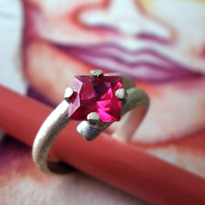 Könnte beinhalten: Ein silberner Ring mit einem quadratischen, leuchtend pinkfarbenen Edelstein, der in der Mitte eingesetzt ist.
