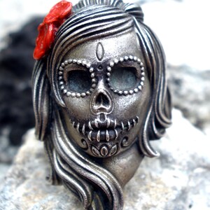 May include: A silver-colored Day of the Dead skull pendant with a red flower accent. The skull features detailed facial markings and long flowing hair. The pendant is designed in a stylized, artistic manner.