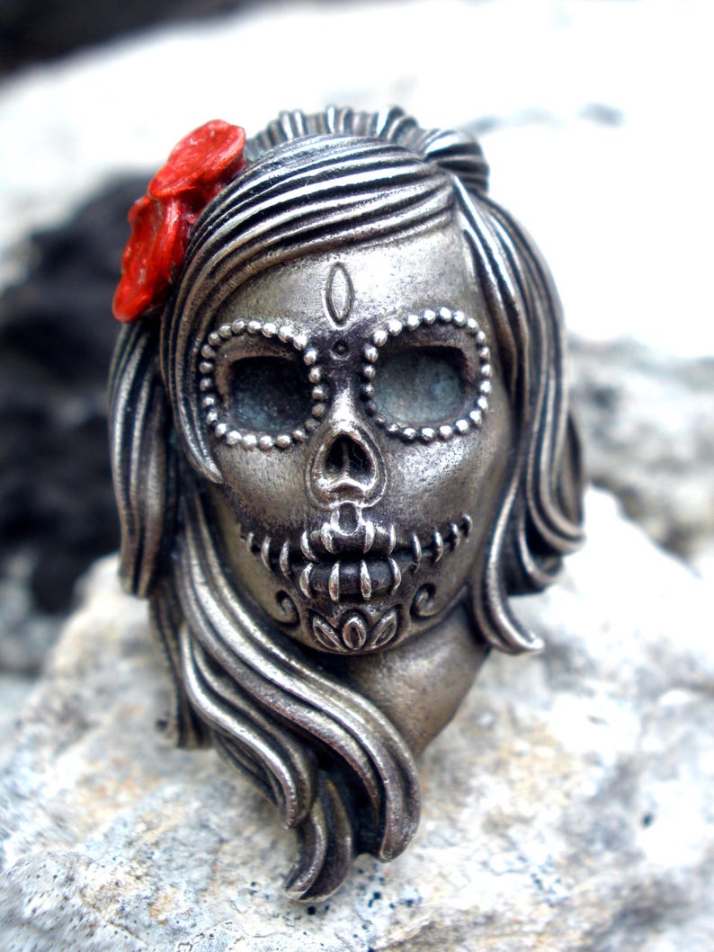 May include: A silver-colored Day of the Dead skull pendant with a red flower accent. The skull features detailed facial markings and long flowing hair. The pendant is designed in a stylized, artistic manner.