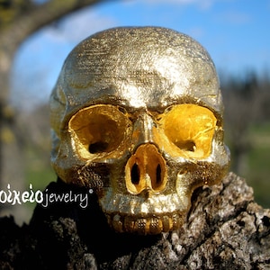 Può includere: Una scultura di teschio dipinta d'oro, con un design dettagliato, si trova su una superficie ruvida di colore marrone. Il teschio è un motivo popolare nei gioielli e nell'arredamento della casa.