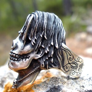 May include: A silver skull ring with detailed features, including a textured hairstyle and a decorative band. The ring is set against a natural, blurred background, highlighting the intricate design of the jewelry.