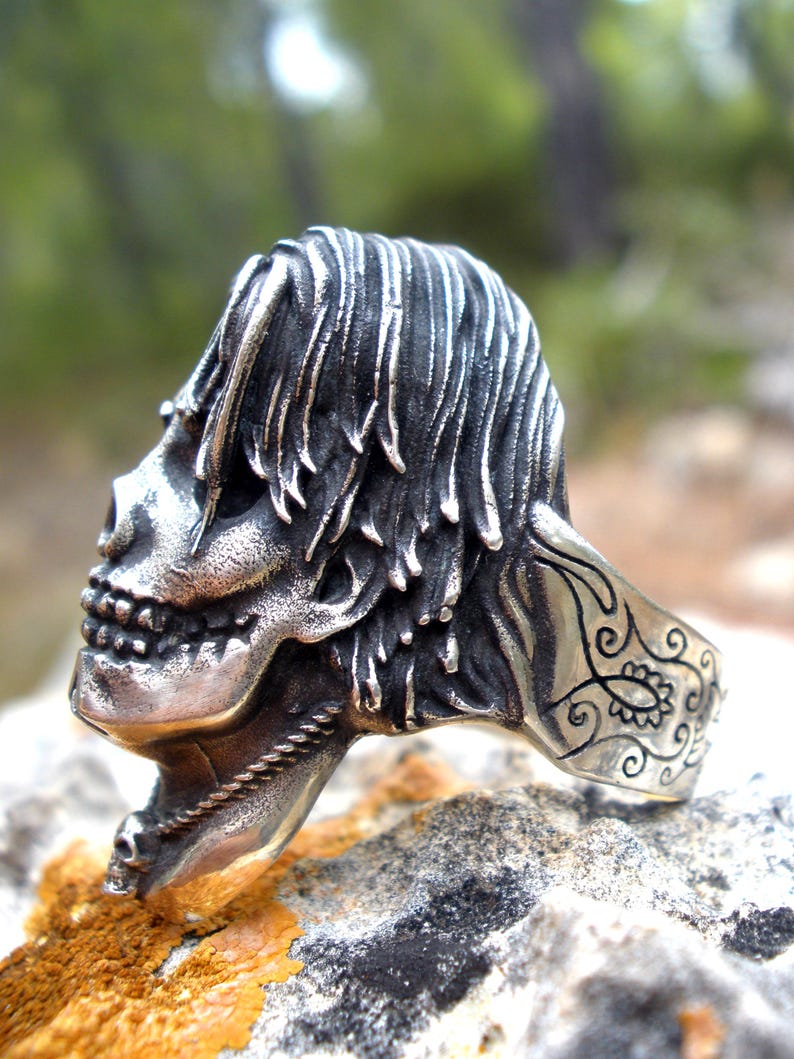 May include: A silver skull ring with detailed features, including a textured hairstyle and a decorative band. The ring is set against a natural, blurred background, highlighting the intricate design of the jewelry.