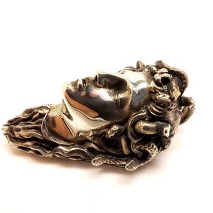 May include: A metallic silver-toned brooch or pendant featuring a sculpted human face in profile. The figure has flowing, textured hair that transitions into intricate, snake-like forms. The surface has a polished, reflective finish on the face.