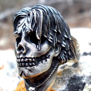 May include: A silver-colored skull ring with long hair and intricate facial details. The ring features a detailed skull design with a necklace and a small skull pendant. The ring is set on a textured surface.