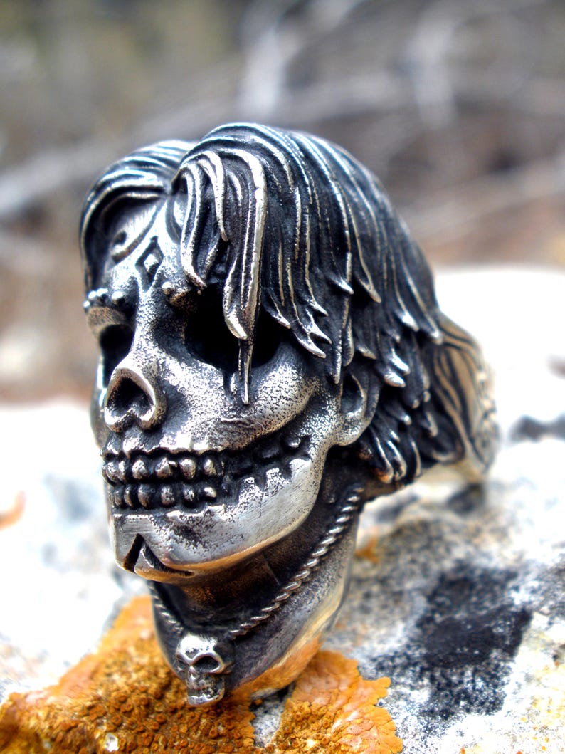 May include: A silver-colored skull ring with long hair and intricate facial details. The ring features a detailed skull design with a necklace and a small skull pendant. The ring is set on a textured surface.