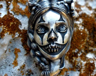 May include: A silver pendant featuring a sculpted face with intricate details, including braided hair, bows, and clown-like makeup with dark eye markings and a stitched mouth. The metal has a polished finish with darkened recessed areas for contrast.
