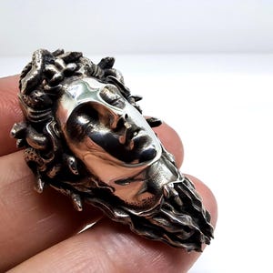 May include: A polished silver ring featuring a detailed sculptural face with closed eyes and flowing hair composed of intertwined snakes. The metal has a high-shine finish on the facial features and a darkened, textured patina on the serpent hair.