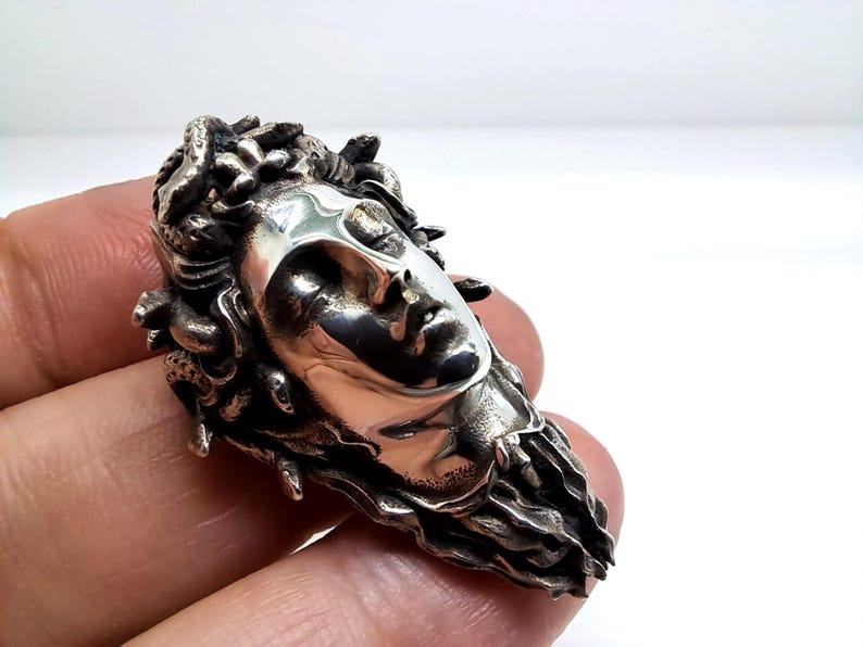 May include: A polished silver ring featuring a detailed sculptural face with closed eyes and flowing hair composed of intertwined snakes. The metal has a high-shine finish on the facial features and a darkened, textured patina on the serpent hair.