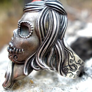 May include: A silver-toned ring featuring a detailed sugar skull design. The skull has intricate facial features, including stitched mouth and beaded eye sockets. The ring's band is decorated with floral patterns and the hair is styled in flowing waves.