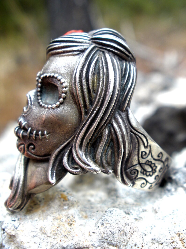 May include: A silver-toned ring featuring a detailed sugar skull design. The skull has intricate facial features, including stitched mouth and beaded eye sockets. The ring's band is decorated with floral patterns and the hair is styled in flowing waves.