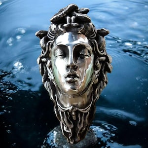 May include: A metallic silver-toned sculpture of a human head with closed eyes and flowing, textured hair. The sculpture features a polished, reflective face contrasted by a dark, oxidized finish on the hair and neck, creating a detailed relief.