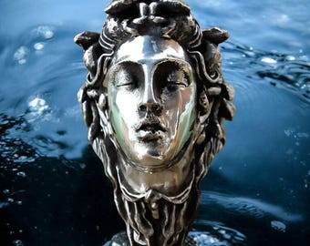 May include: A metallic silver-toned sculpture of a human head with closed eyes and flowing, textured hair. The sculpture features a polished, reflective face contrasted by a dark, oxidized finish on the hair and neck, creating a detailed relief.