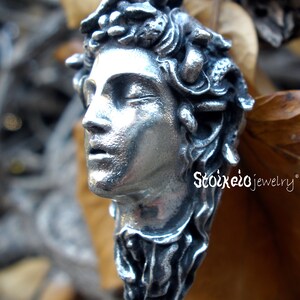 May include: A silver-toned pendant shaped like a human head with snakes for hair, featuring closed eyes and parted lips. The piece has a textured, metallic finish and is suspended from a black cord. The text Stoikeio jewelry is visible.