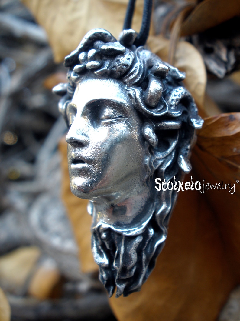 May include: A silver-toned pendant shaped like a human head with snakes for hair, featuring closed eyes and parted lips. The piece has a textured, metallic finish and is suspended from a black cord. The text Stoikeio jewelry is visible.