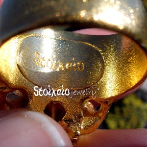 May include: A gold-toned ring with a textured, brushed finish on the inner band. The interior of the band features the engraved text Stoixeio. The text Stoixeio jewelry is also printed in white across the center of the ring's surface.