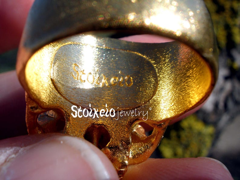 May include: A gold-toned ring with a textured, brushed finish on the inner band. The interior of the band features the engraved text Stoixeio. The text Stoixeio jewelry is also printed in white across the center of the ring's surface.