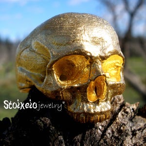 May include: A metallic gold-colored ring shaped like a human skull with detailed facial features including eye sockets, a nasal cavity, and teeth. The text Stoixeiojewelry is printed in white on the lower left side of the skull ring.