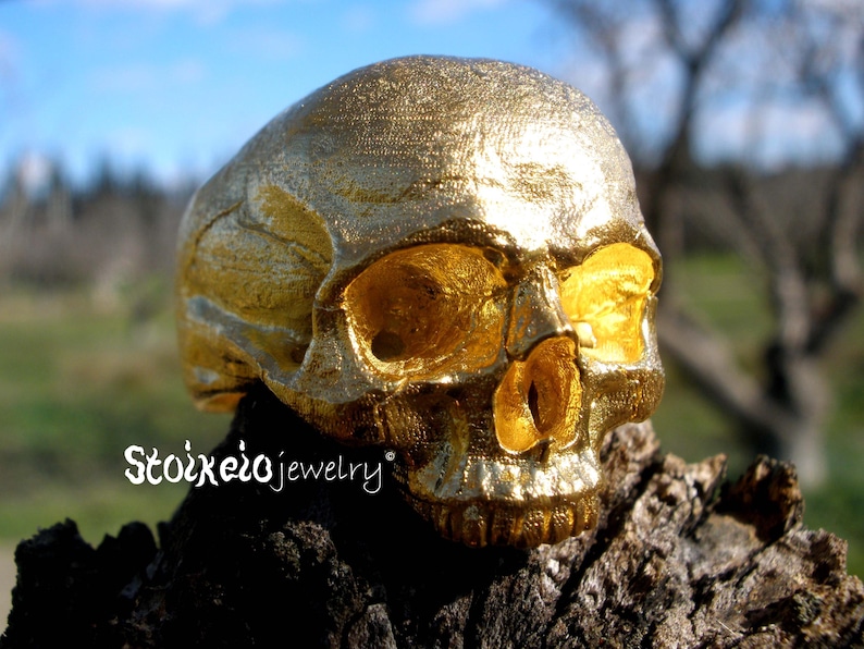 May include: A metallic gold-colored ring shaped like a human skull with detailed facial features including eye sockets, a nasal cavity, and teeth. The text Stoixeiojewelry is printed in white on the lower left side of the skull ring.