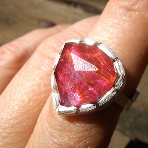 Op de afbeelding: Een zilveren ring met een grote, gefacetteerde, roze edelsteen. De edelsteen heeft een uniek, gestreept patroon.