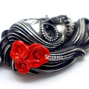 May include: A silver-toned pendant in the shape of a woman's face with flowing hair, adorned with three bright red roses. The pendant has intricate details and the text "Stoixelo jewelry".