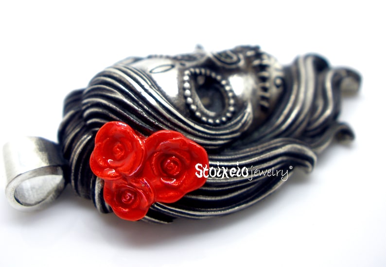 May include: A silver-toned pendant in the shape of a woman's face with flowing hair, adorned with three bright red roses. The pendant has intricate details and the text "Stoixelo jewelry".