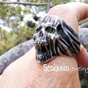 May include: A silver-toned metal ring featuring a detailed, weathered skull design with an open mouth and draped fabric-like textures on the sides. The text Stoixeio jewelry is printed in white in the lower right corner of the image.