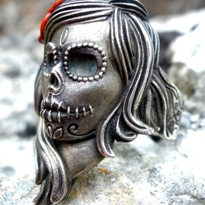 May include: A silver-toned ring in the shape of a sugar skull, a Day of the Dead motif. The skull has detailed features, including stitched mouth, beaded eye sockets, and flowing hair. A small red accent is visible on the top of the head.