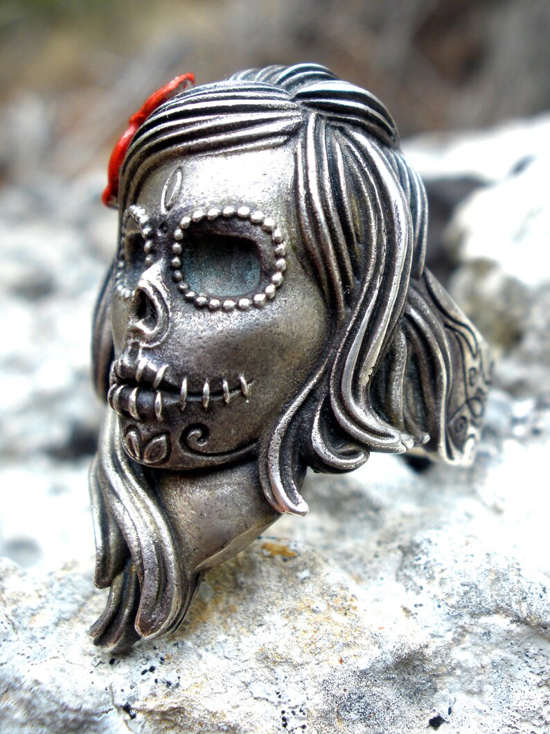 May include: A silver-toned ring in the shape of a sugar skull, a Day of the Dead motif. The skull has detailed features, including stitched mouth, beaded eye sockets, and flowing hair. A small red accent is visible on the top of the head.