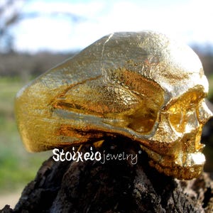 May include: A gold-colored ring shaped like a human skull with a textured, metallic finish. The side of the ring features the text Stoixeio jewelry in a white script font. The skull design covers the entire band, showing detailed facial features.