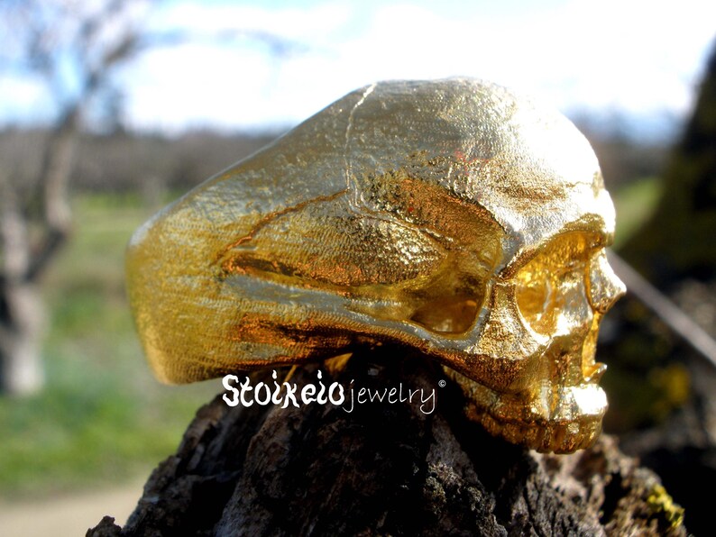 May include: A gold-colored ring shaped like a human skull with a textured, metallic finish. The side of the ring features the text Stoixeio jewelry in a white script font. The skull design covers the entire band, showing detailed facial features.