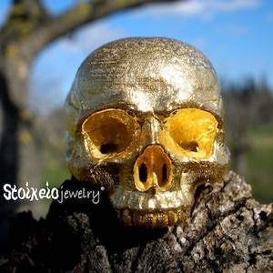 May include: A metallic gold-colored skull sculpture with a textured surface and hollow eye sockets. The text Stoixeio jewelry is printed in white on the left side of the skull. The object features detailed bone structure and a reflective finish.