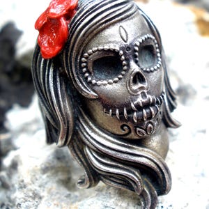 May include: A silver-toned Day of the Dead skull pendant with flowing hair and a red flower. The skull features detailed facial markings and is a unique piece of jewelry.