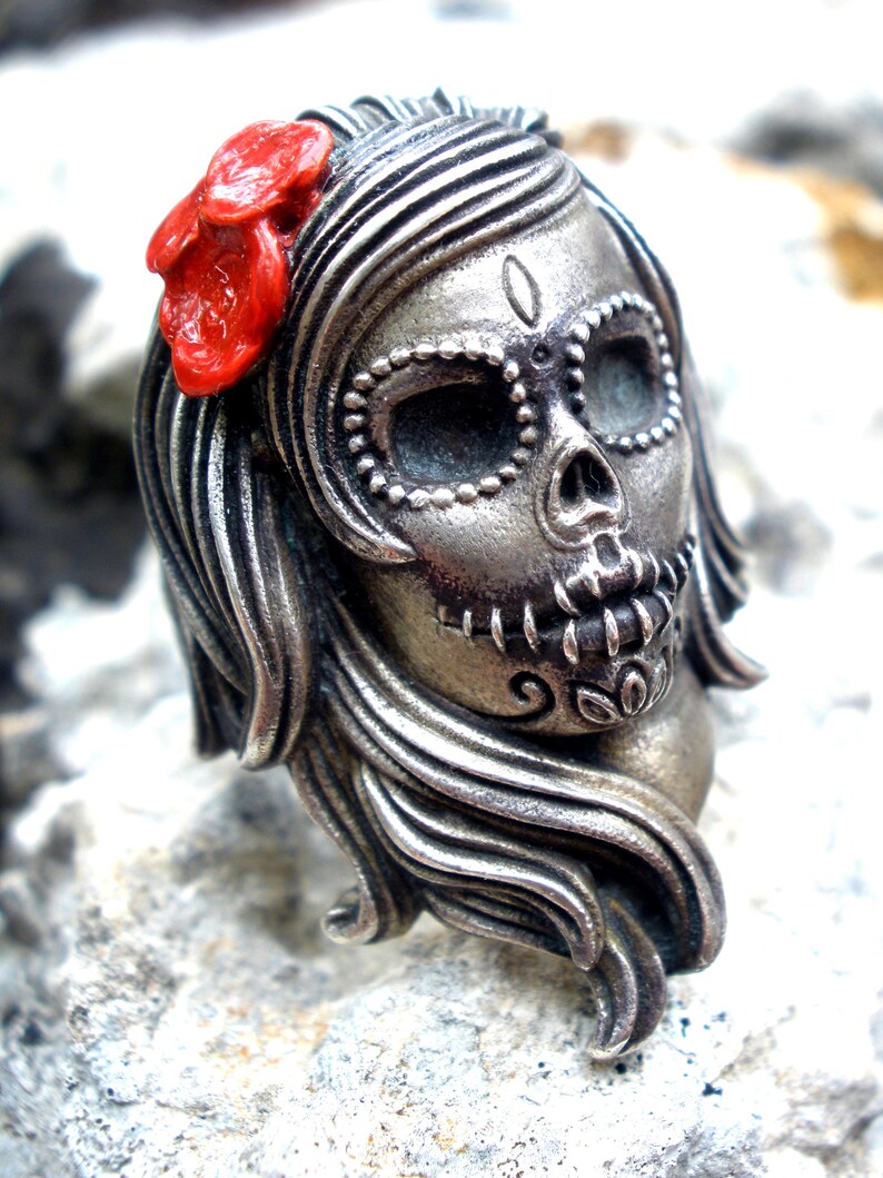 May include: A silver-toned Day of the Dead skull pendant with flowing hair and a red flower. The skull features detailed facial markings and is a unique piece of jewelry.