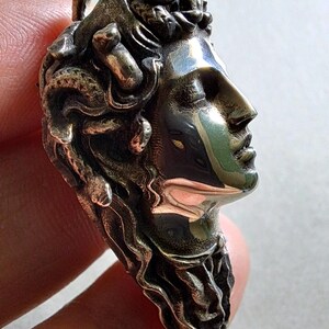 May include: A metallic pendant featuring a sculpted profile of a face with closed eyes and hair composed of intertwined snakes. The surface has a polished, reflective finish with dark, recessed areas that highlight the intricate details of the design.