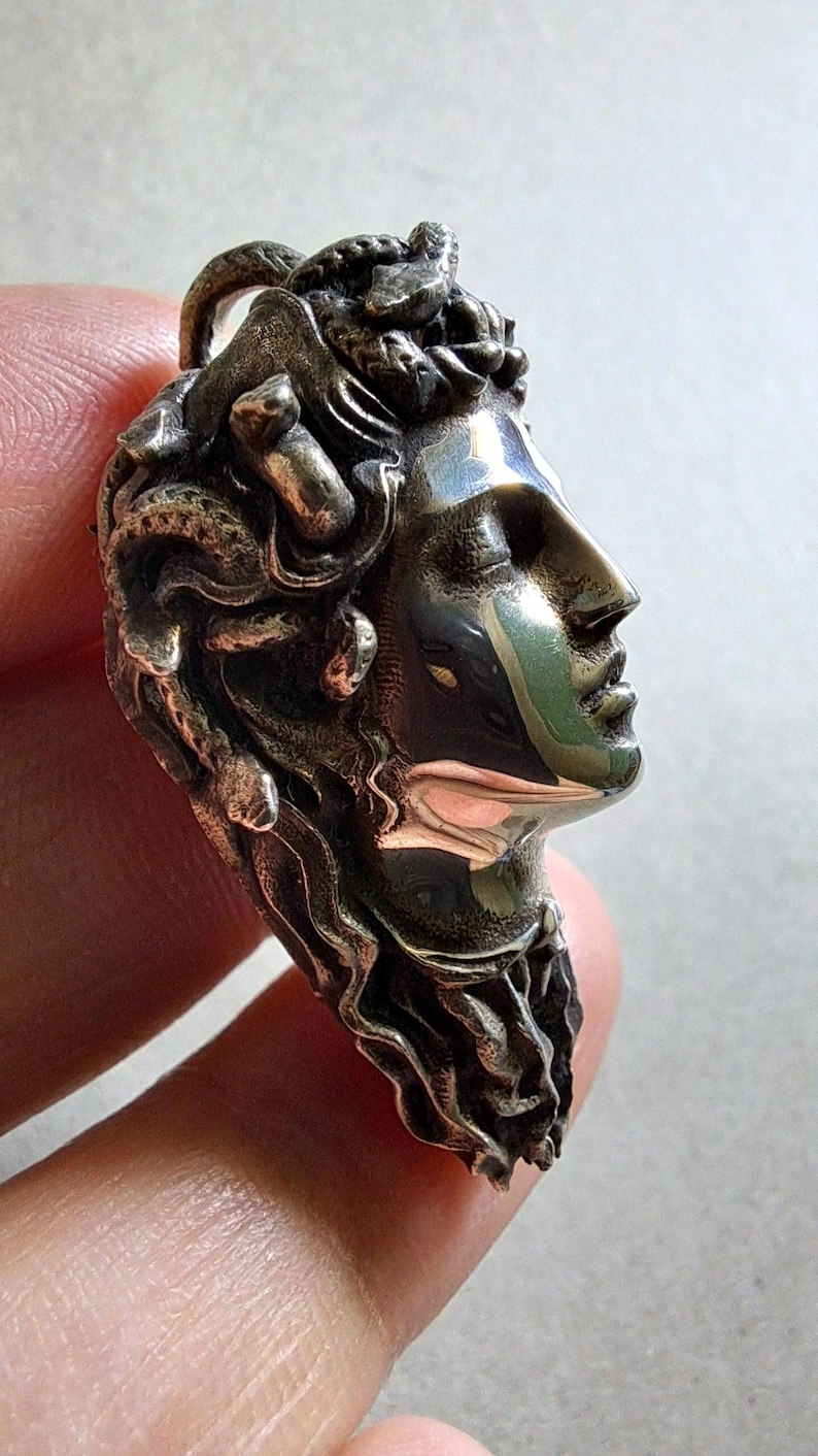 May include: A metallic pendant featuring a sculpted profile of a face with closed eyes and hair composed of intertwined snakes. The surface has a polished, reflective finish with dark, recessed areas that highlight the intricate details of the design.