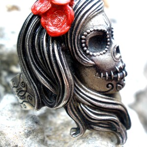 May include: A silver-toned ring in the shape of a sugar skull, adorned with flowing hair and a red flower. The ring features intricate details, including beaded accents around the eye sockets and stitched mouth details. The ring is set against a textured background.