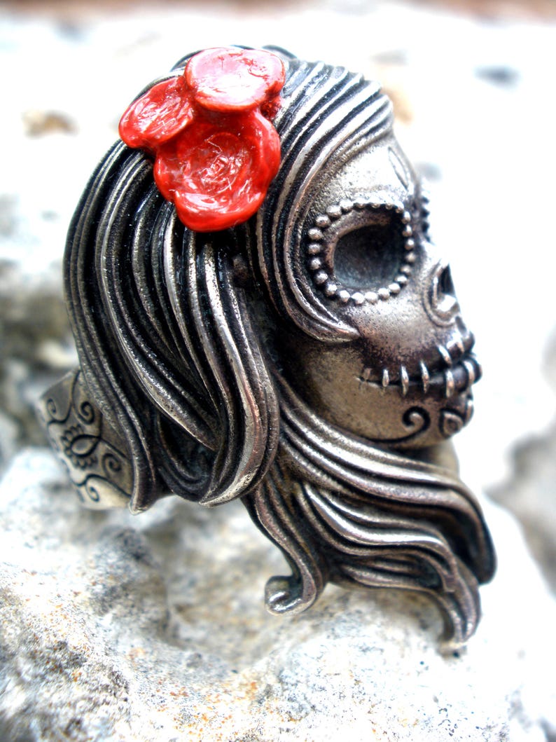 May include: A silver-toned ring in the shape of a sugar skull, adorned with flowing hair and a red flower. The ring features intricate details, including beaded accents around the eye sockets and stitched mouth details. The ring is set against a textured background.