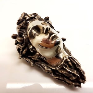 May include: A metallic brooch featuring a sculpted face with closed eyes, surrounded by textured, snake-like forms. The central face has a smooth, polished finish, while the surrounding hair and serpent details have a dark, oxidized metal appearance.