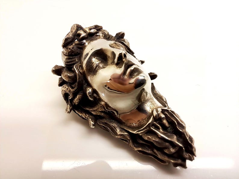 May include: A metallic brooch featuring a sculpted face with closed eyes, surrounded by textured, snake-like forms. The central face has a smooth, polished finish, while the surrounding hair and serpent details have a dark, oxidized metal appearance.