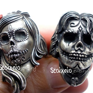 May include: Two silver-toned rings, each sculpted in the shape of a sugar skull. One ring features a female skull with a red bow, the other a male skull. Both rings have intricate details and are from Stoixeio jewelry.