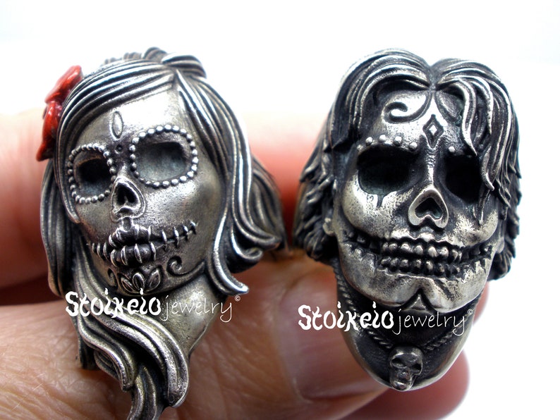 May include: Two silver-toned rings, each sculpted in the shape of a sugar skull. One ring features a female skull with a red bow, the other a male skull. Both rings have intricate details and are from Stoixeio jewelry.
