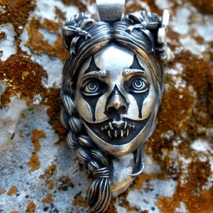 May include: A silver pendant featuring a sculpted face with intricate details, including braided hair, bows, and dark markings around the eyes and mouth. The design includes a stitched mouth effect and a bail at the top for hanging on a chain.